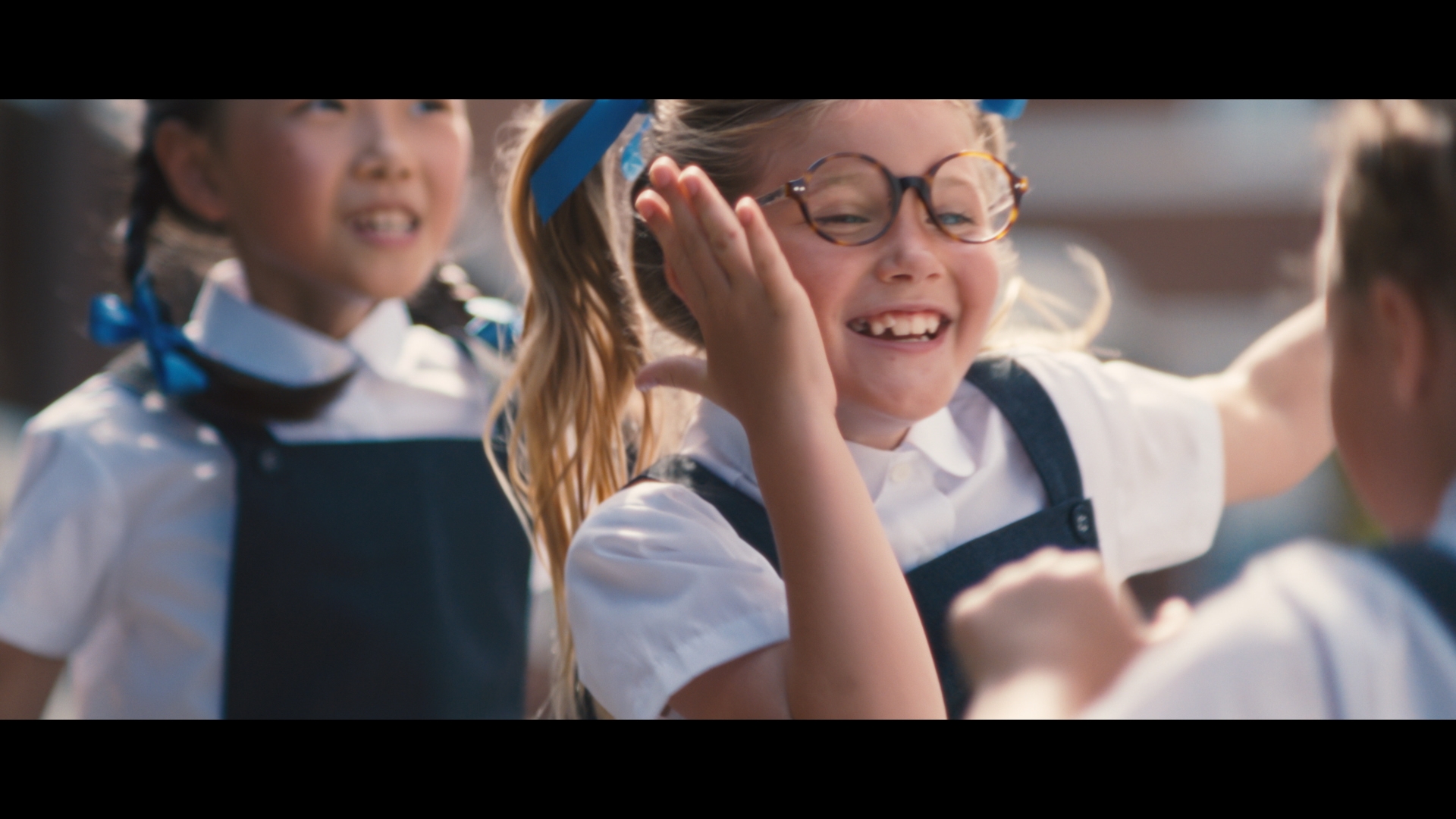 Group of children in school uniforms outdoors. A blond girl with glasses smiles broadly among her classmates on a sunny day.