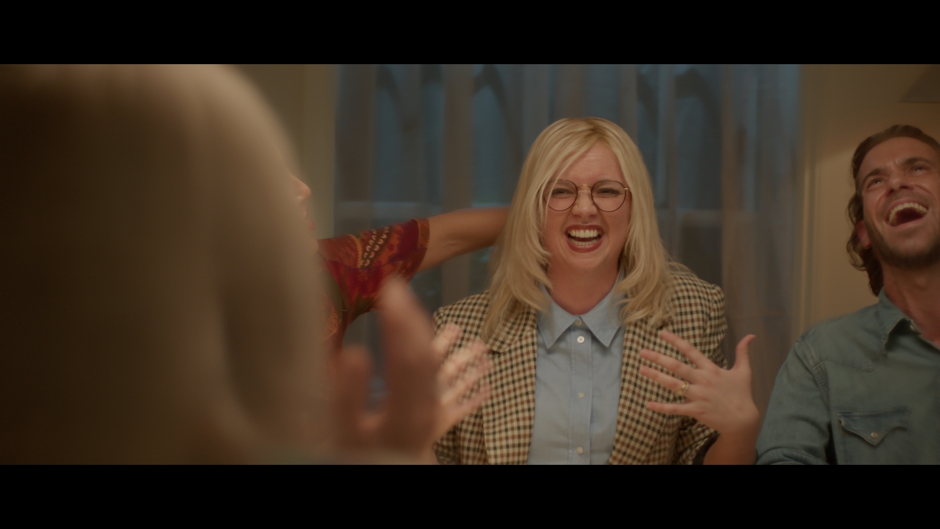 Group of friends laughing and gesturing around a table indoors, with one woman in a blazer and glasses as a focal point.