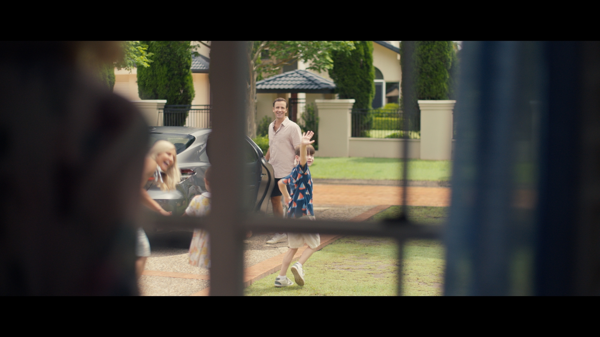 Father and children near a grey car parked outside of modern home with lush green lawn and manicured bushes on a sunny day.