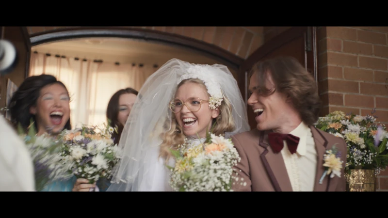 Bride and groom and wedding party celebrate and laugh outside a brick building, bouquets and smiles on a bright day.