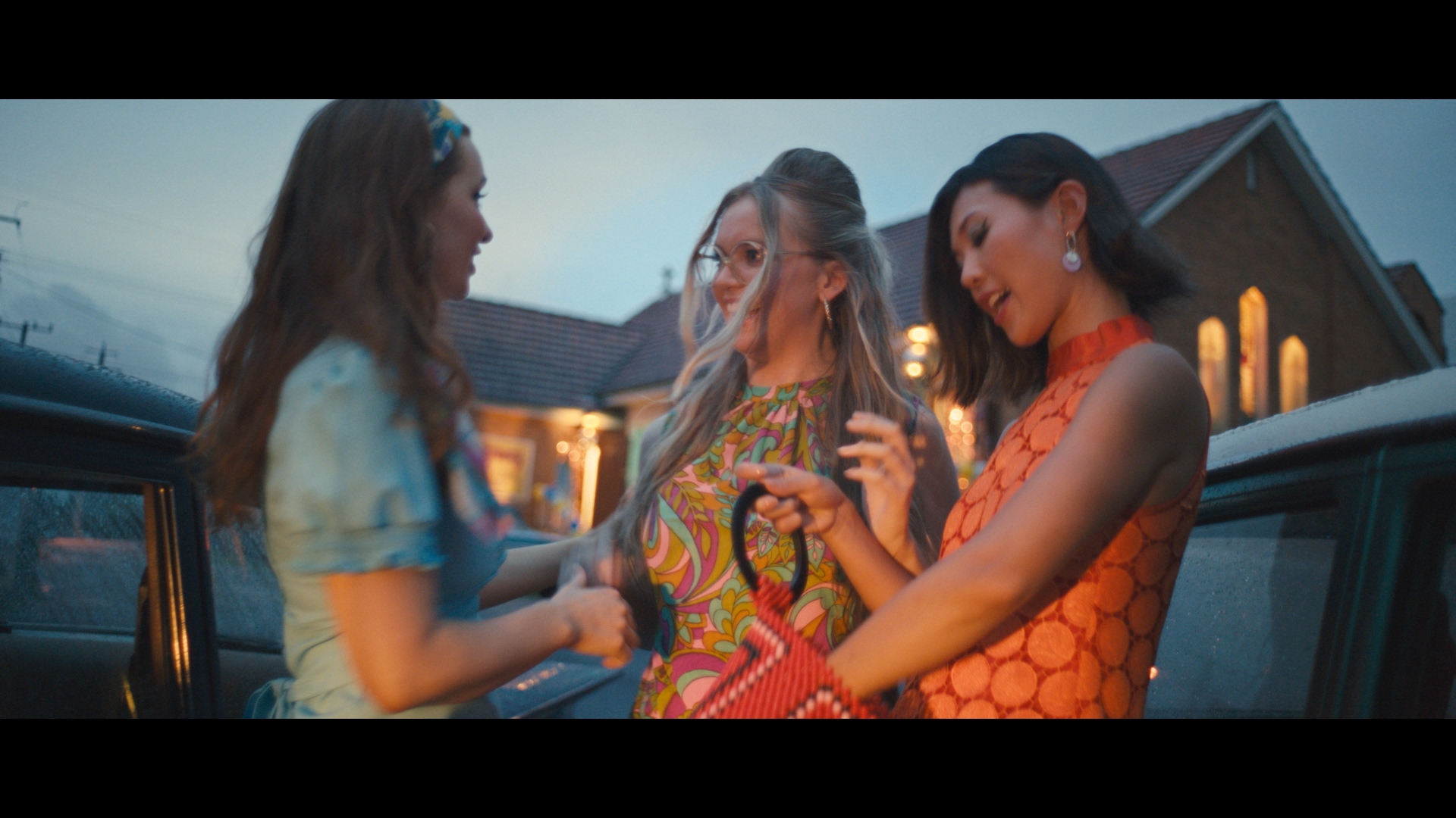 Three young women in 1960s-inspired dresses standing near a car at dusk, with a retro aesthetic and a brick building in the background.