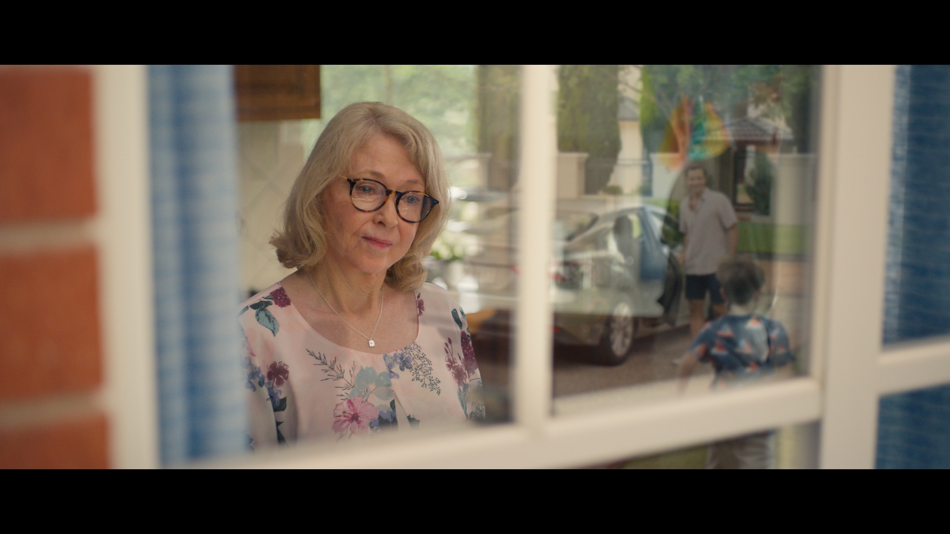 Older woman with glasses looking through a window with reflection of man and child outside in a residential setting during daytime.