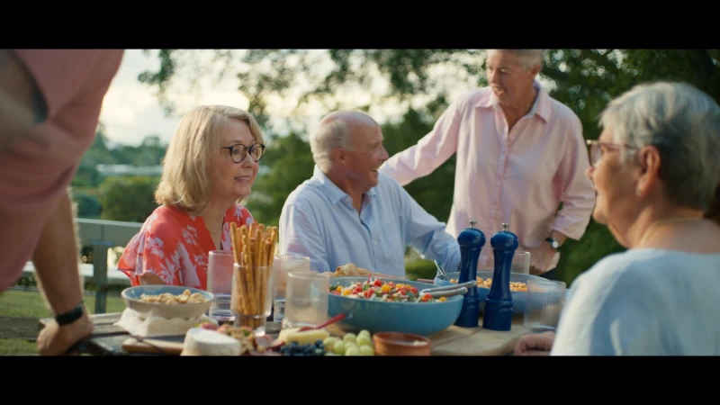 Outdoor shot of a group of older adults gathered around a table with food and drinks, enjoying conversation and each other's company.
