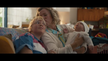An older woman kissing a younger woman on the cheek while she holds a crying baby on a sofa in a living room.