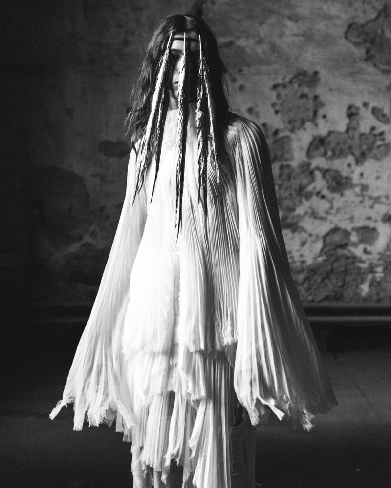 Monochrome studio shot of a solitary adult in draped white fabric, long dark hair with light streaks, and a distressed gray wall.
