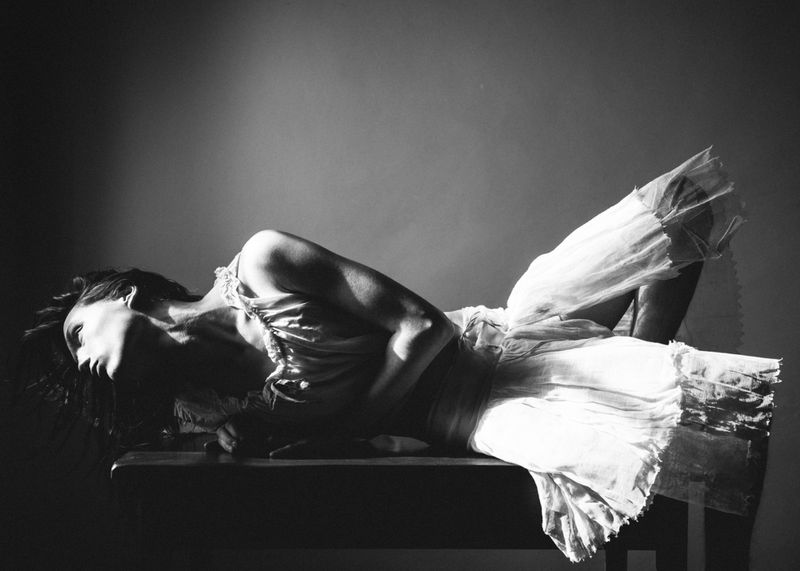 Black and white studio shot of a female figure in motion, wearing a flowing white dress, laying on a dark surface in dramatic lighting.