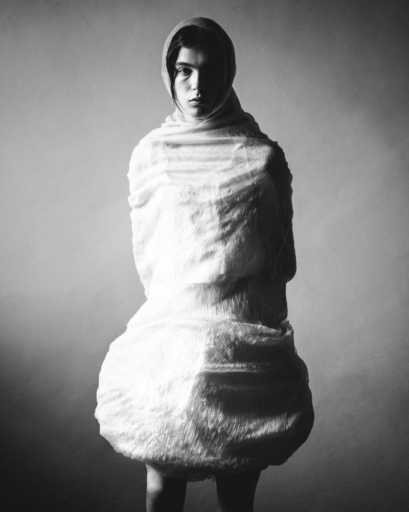 Woman in monochrome wrapped in textured white fabric with a hood, standing in a studio with soft lighting, fashion editorial photography.