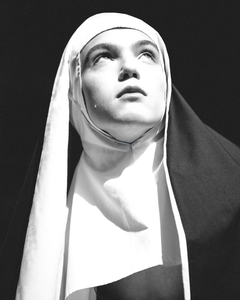 Monochrome portrait of a woman wearing a nun's habit, looking upwards with a tear on her cheek, against a dark background.