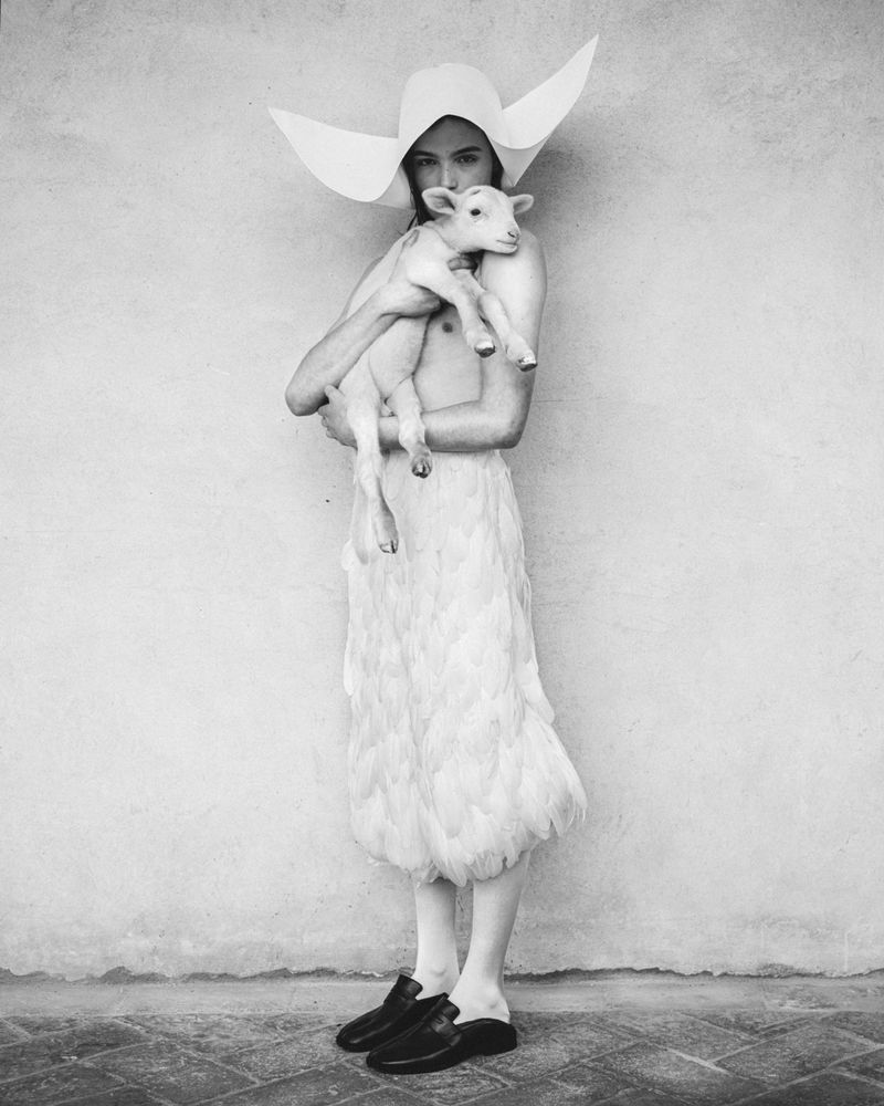 Monochrome portrait of a person holding a lamb, wearing a wide-brimmed hat and feathered skirt against a textured grey wall backdrop.