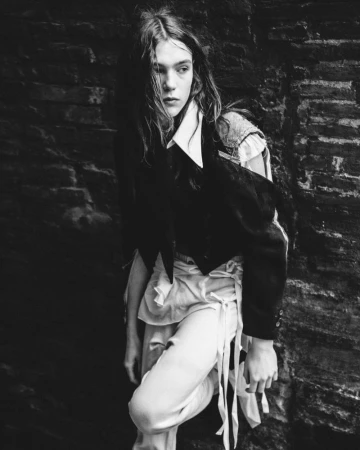 A person with long hair in a black vest and white pants poses against a textured dark background in a black and white fashion image.