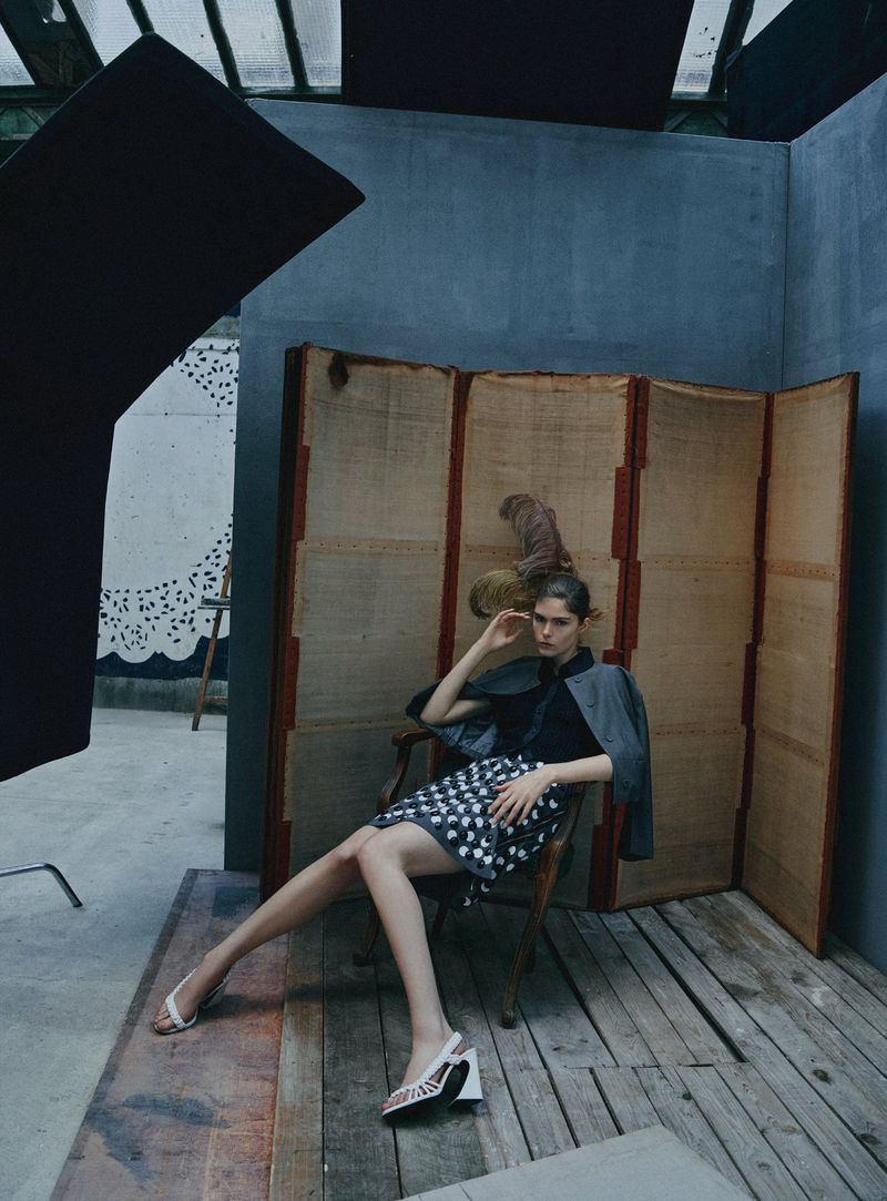 Model Anna Robinson sits in a wooden armchair before a tall folding screen wearing a patterned skirt and white heels.