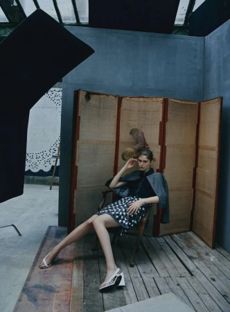 Model Anna Robinson sits in a wooden armchair before a tall folding screen wearing a patterned skirt and white heels.