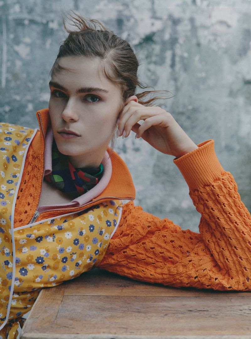 Model Anna Robinson wearing an orange knit sweater and floral print vest leaning on a wooden table against a textured wall.
