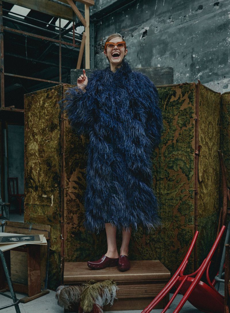 Adult woman in a blue faux fur coat and orange eyeglasses strikes a pose indoors. She stands on a wooden platform in a textured room.