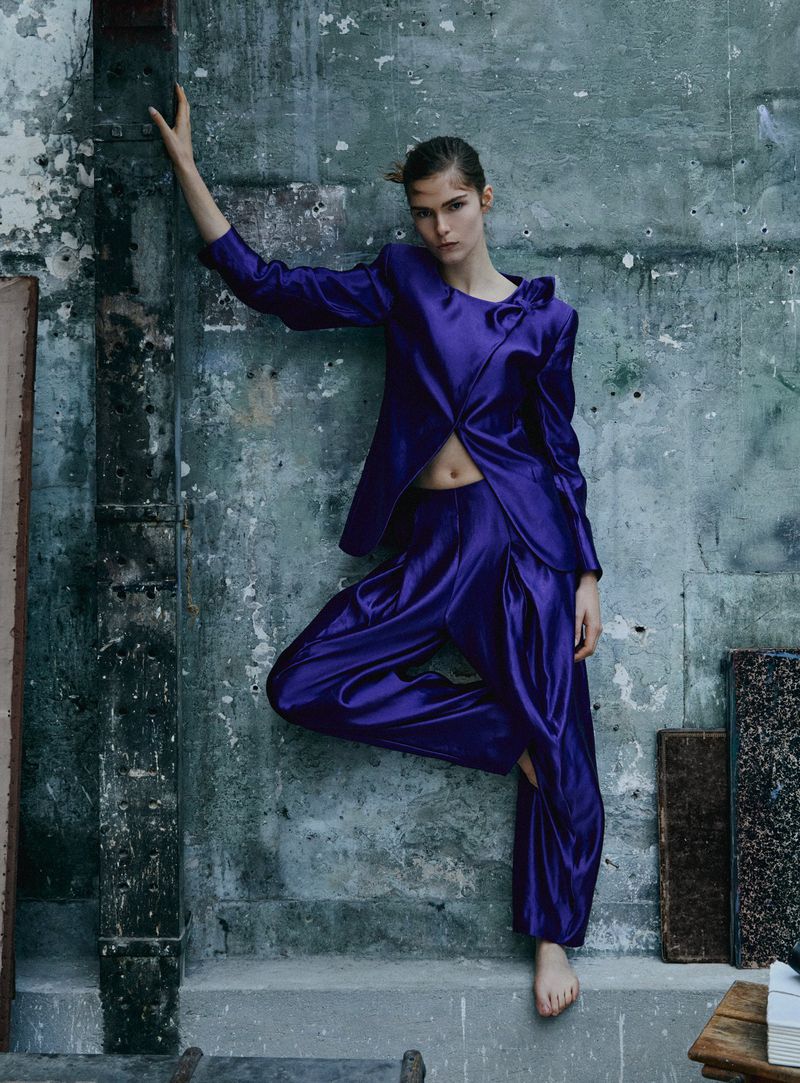 Anna Robinson wearing a vibrant purple satin suit and posing barefoot against a distressed concrete wall.