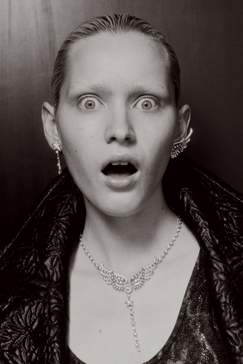 Close-up of a blonde adult woman with surprised expression, adorned with a sparkling necklace and earrings against a dark backdrop.