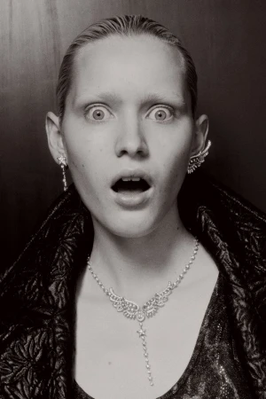 Close-up of a blonde adult woman with surprised expression, adorned with a sparkling necklace and earrings against a dark backdrop.