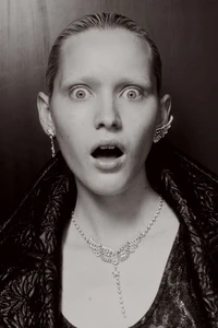 Close-up portrait of a woman with slicked-back hair and a surprised expression wearing elaborate silver jewelry and a dark quilted jacket.