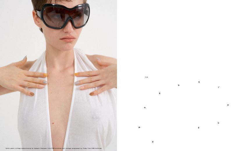 Model Greta Hofer wearing oversized black visor sunglasses and a plunging white halter garment against a clean white background.