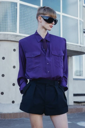 Model wearing black wrap-around sunglasses, a structured purple button-down shirt, necktie, and black tailored pleated shorts.