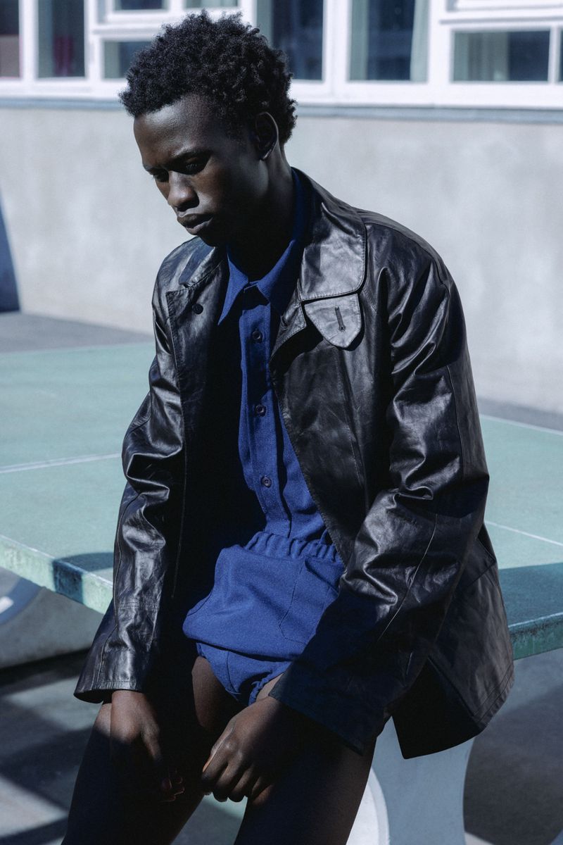 Young Black male model wearing a black leather jacket and blue shirt seated outdoors for Numero Netherlands.