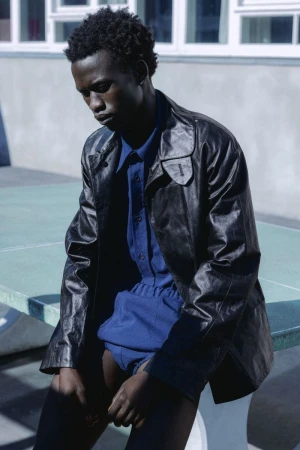 Young Black male model wearing a black leather jacket and blue shirt seated outdoors for Numero Netherlands.