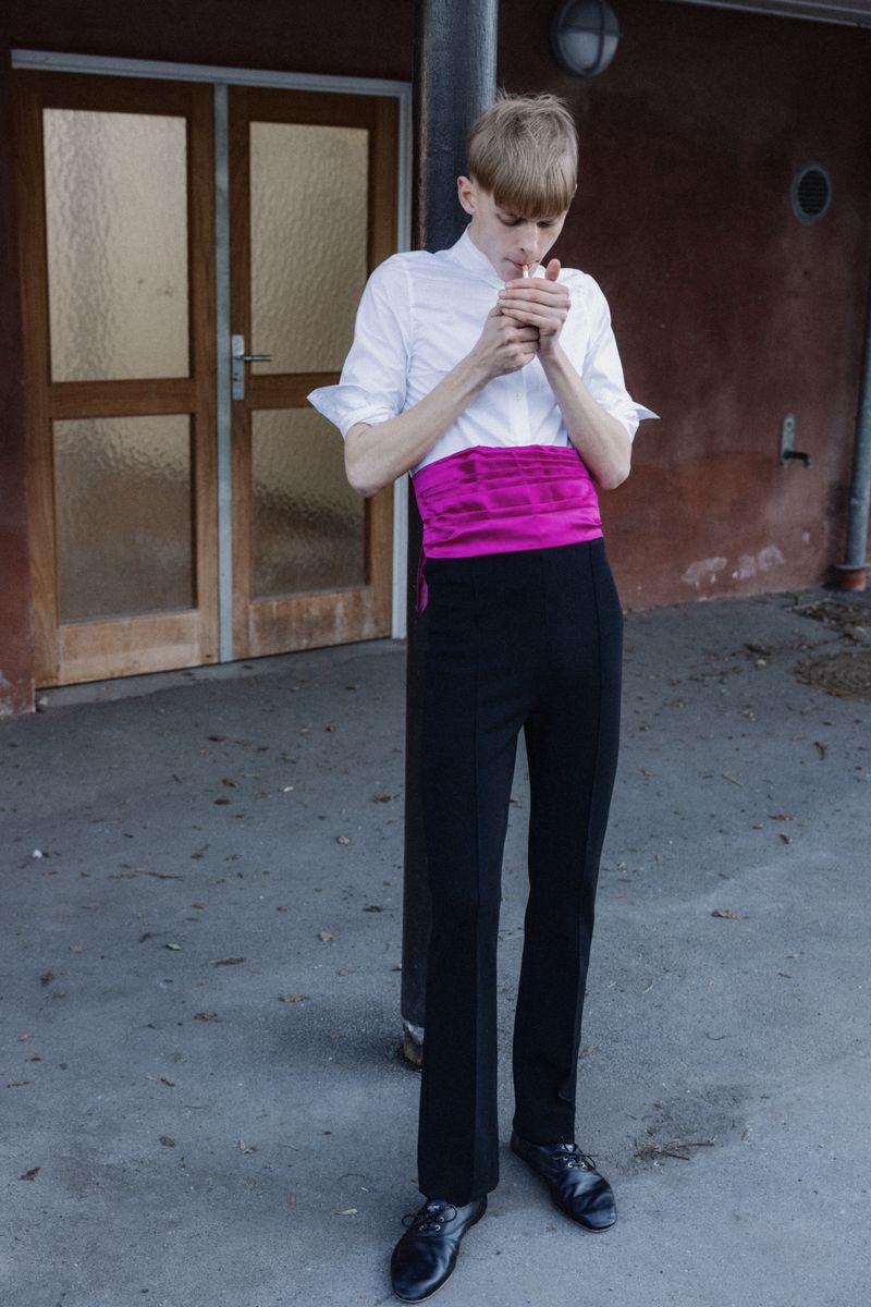 Fashion model in a white shirt, magenta sash, and black trousers lighting a cigarette outdoors by a doorway.