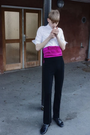 Fashion model in a white shirt, magenta sash, and black trousers lighting a cigarette outdoors by a doorway.