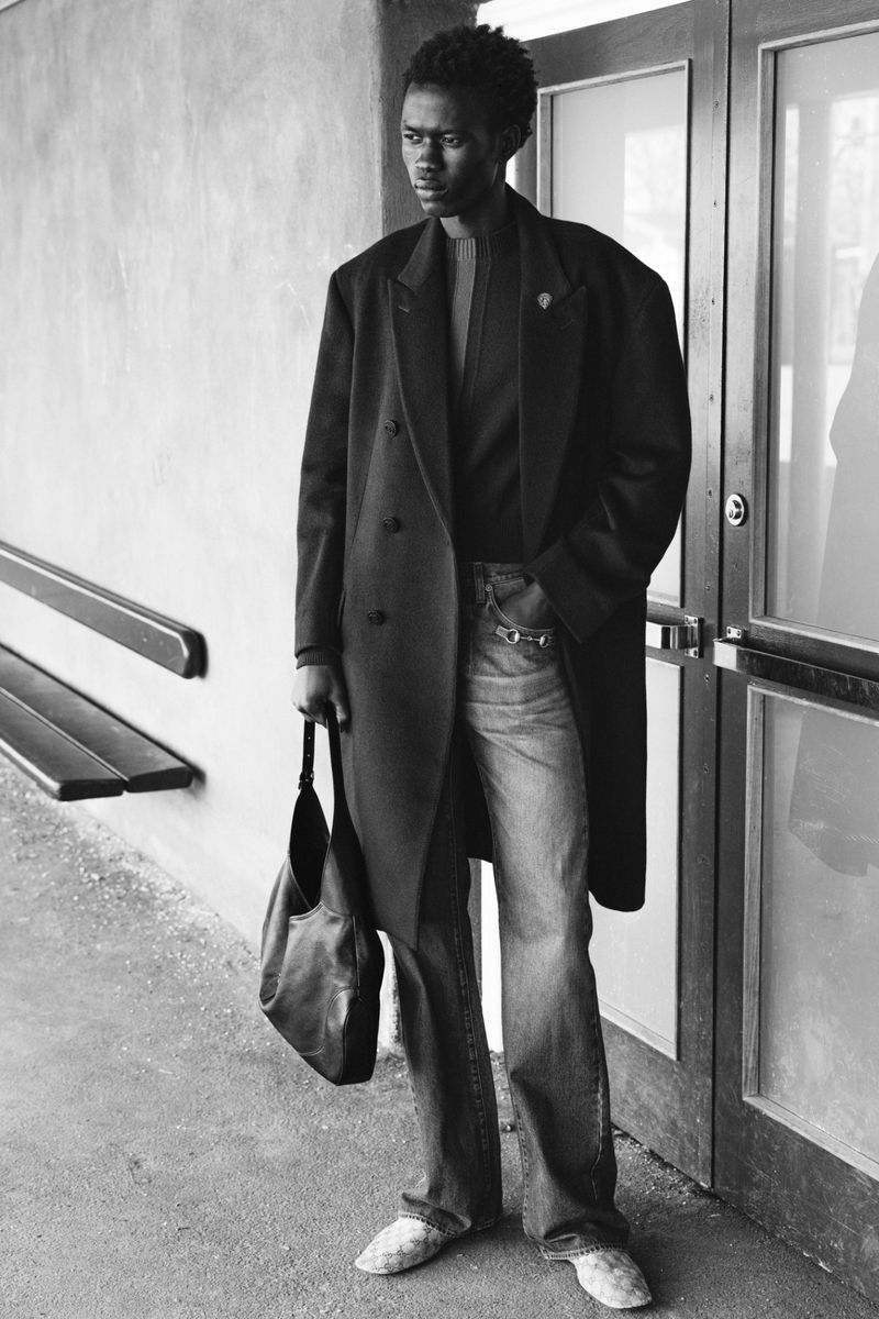 Black male model in an oversized dark coat and wide-leg jeans holding a black leather tote bag beside glass framed doors.