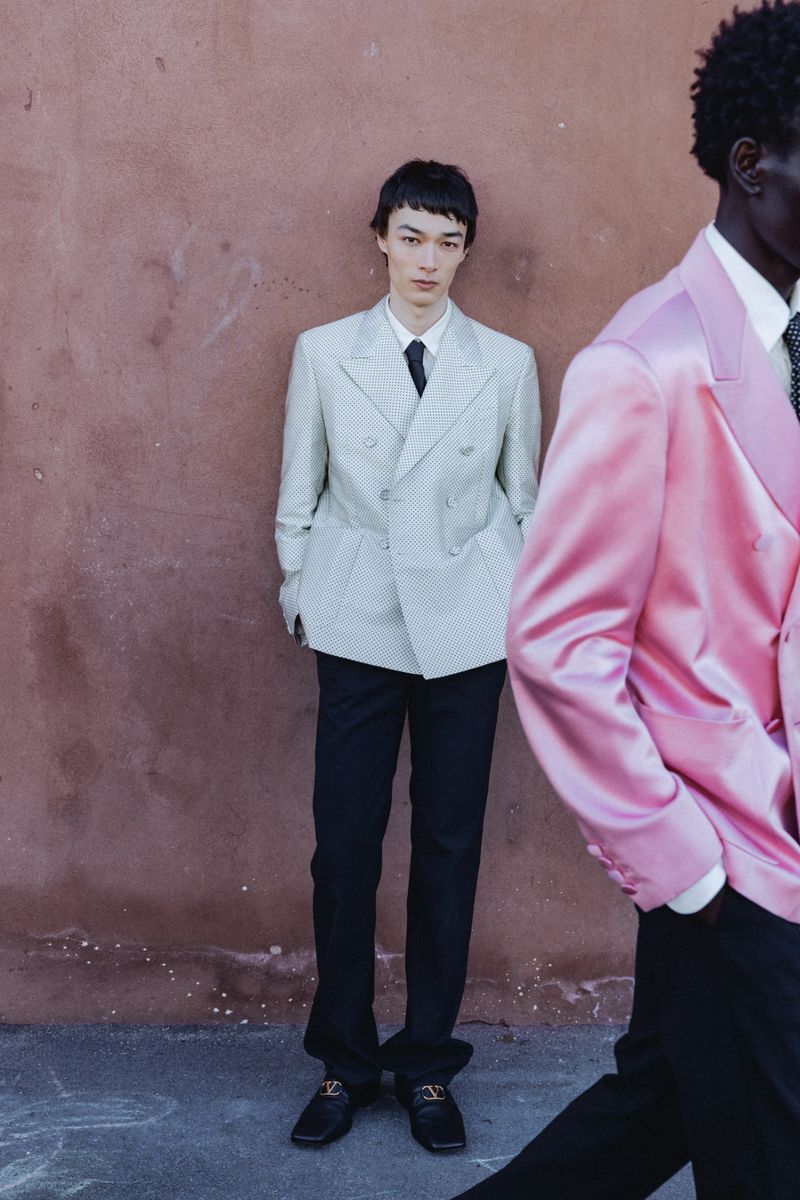 Model in an off-white double-breasted blazer stands by a wall while another model walks past in a shiny pink satin suit jacket.