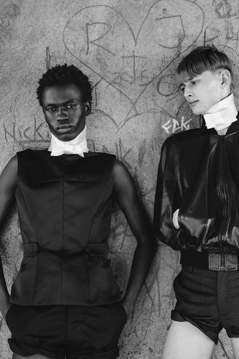 Two male models posing against a graffiti-scratched concrete wall in black and white tailored garments with white neckpieces.