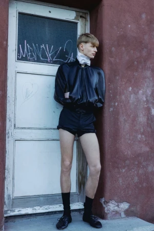 Young male model in black bomber jacket and shorts leaning against weathered pink doorway for Numero Netherlands.