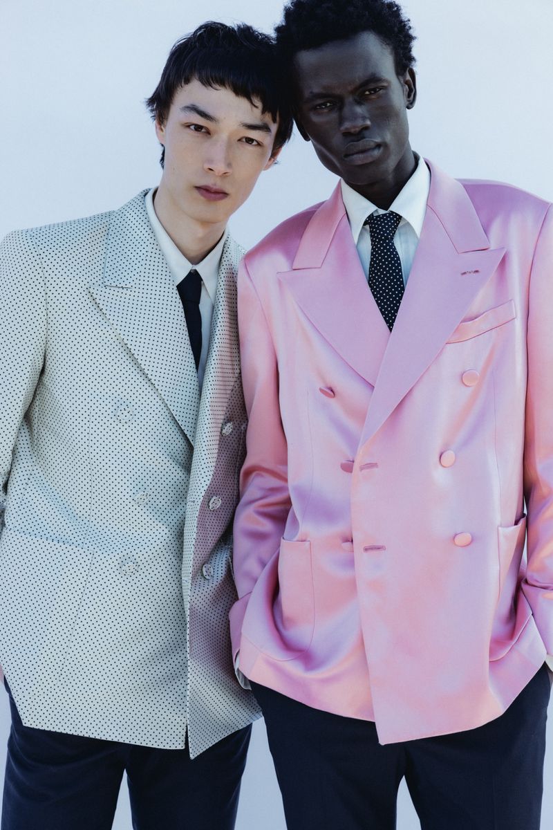 Two male models posing in double-breasted suits, one in light textured grey and one in pink satin, against a white studio background.