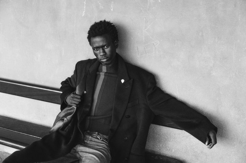 Black man wearing an oversized dark coat and knit sweater seated on a bench leaning against a concrete wall in black and white.