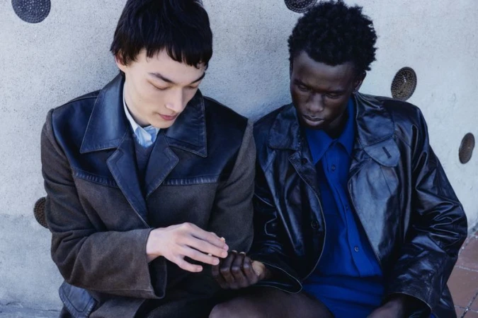 Two male models seated together in leather outerwear and collared shirts against a textured concrete wall.