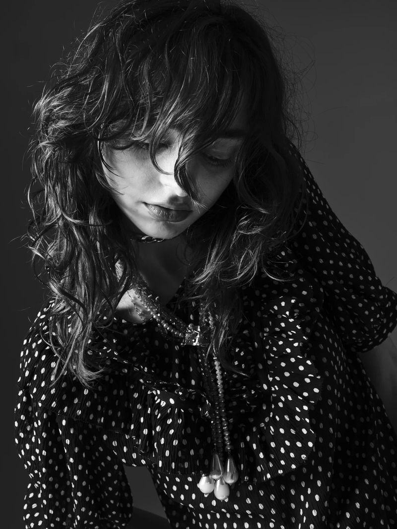 Monochrome portrait of a woman with long curly hair wearing a polka-dot top and layered necklaces, with a muted gray background.