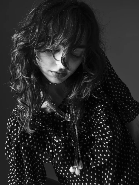 Monochrome portrait of a woman with long curly hair wearing a polka-dot top and layered necklaces, with a muted gray background.