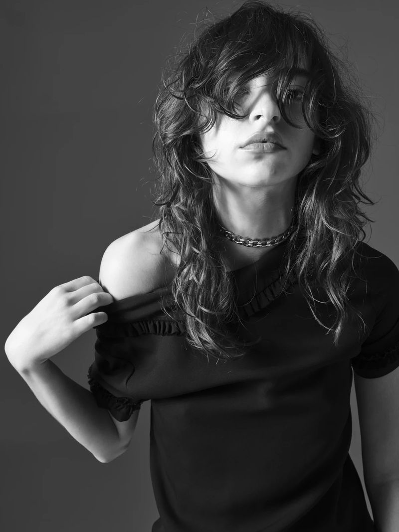 Black and white portrait of a person with long, wavy dark hair, wearing a black top and silver chain necklace, against a gray backdrop.
