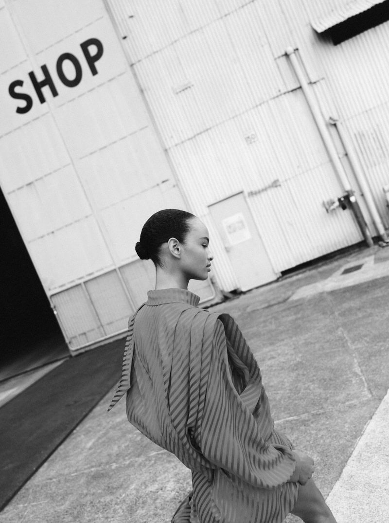 Monochrome fashion shot of a woman in a striped top walking by an industrial building with text 'SHOP' on the side.