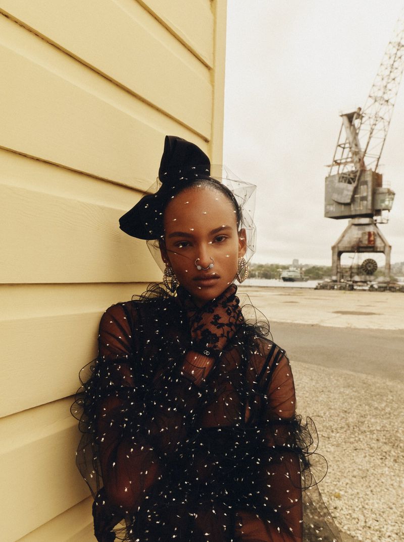 Fashion editorial shot of an adult female wearing a black dress with a black bow, leaning against a yellow wall with a harbor crane.