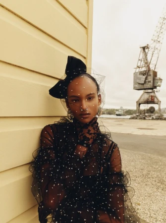 Fashion editorial shot of an adult female wearing a black dress with a black bow, leaning against a yellow wall with a harbor crane.
