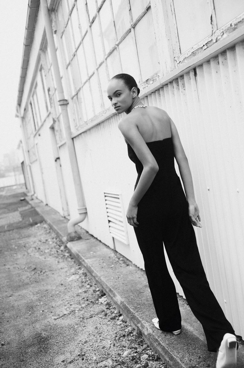 Black and white portrait of a woman in a jumpsuit standing outside a building with multiple windows looking over her shoulder.