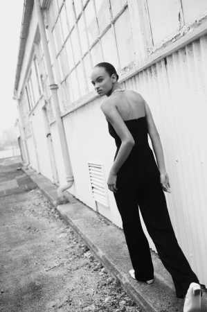 Black and white portrait of a woman in a jumpsuit standing outside a building with multiple windows looking over her shoulder.