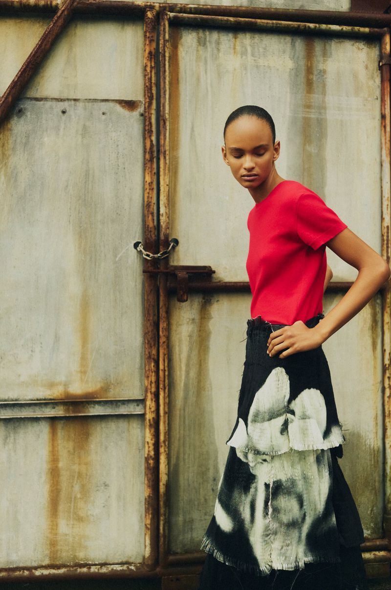 Woman poses in red top and black-and-white skirt against a distressed metal background. Fashion editorial style, outdoor setting.