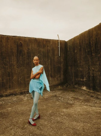 Full-length portrait of a slender African woman in baby blue fashion apparel and red Mary Jane shoes against a concrete wall outdoors.