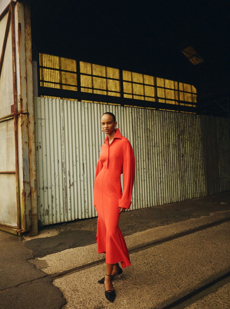 A young woman poses outdoors wearing a red dress and black heels in front of an industrial metal building with yellow windows.