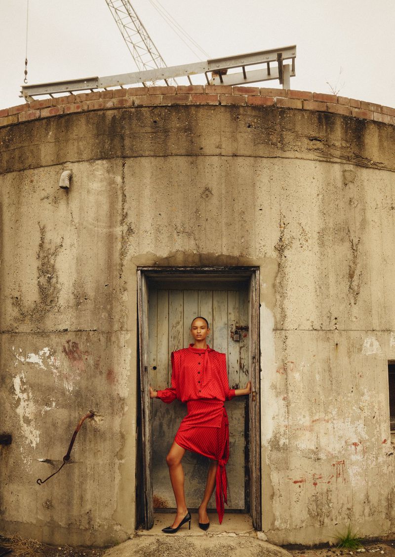 Fashion shot of woman in red dress posing in doorway of concrete structure with crane in background, against overcast sky.