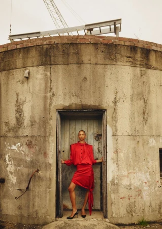 Fashion shot of woman in red dress posing in doorway of concrete structure with crane in background, against overcast sky.