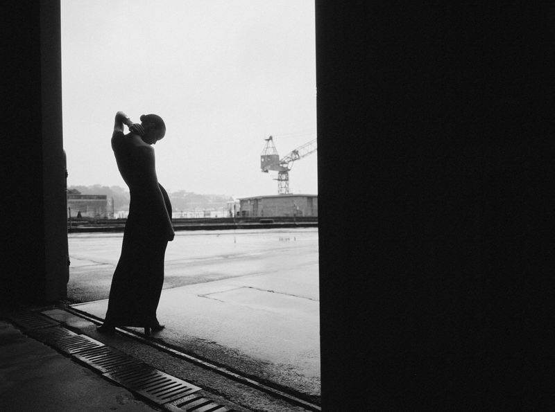 Woman in a long dress poses in silhouette against a waterfront industrial setting with cranes, creating a dramatic fashion image.