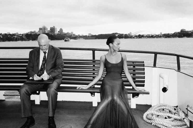 Black and white shot of an adult male in a suit and an adult female in an evening gown sitting on a boat with a city backdrop.
